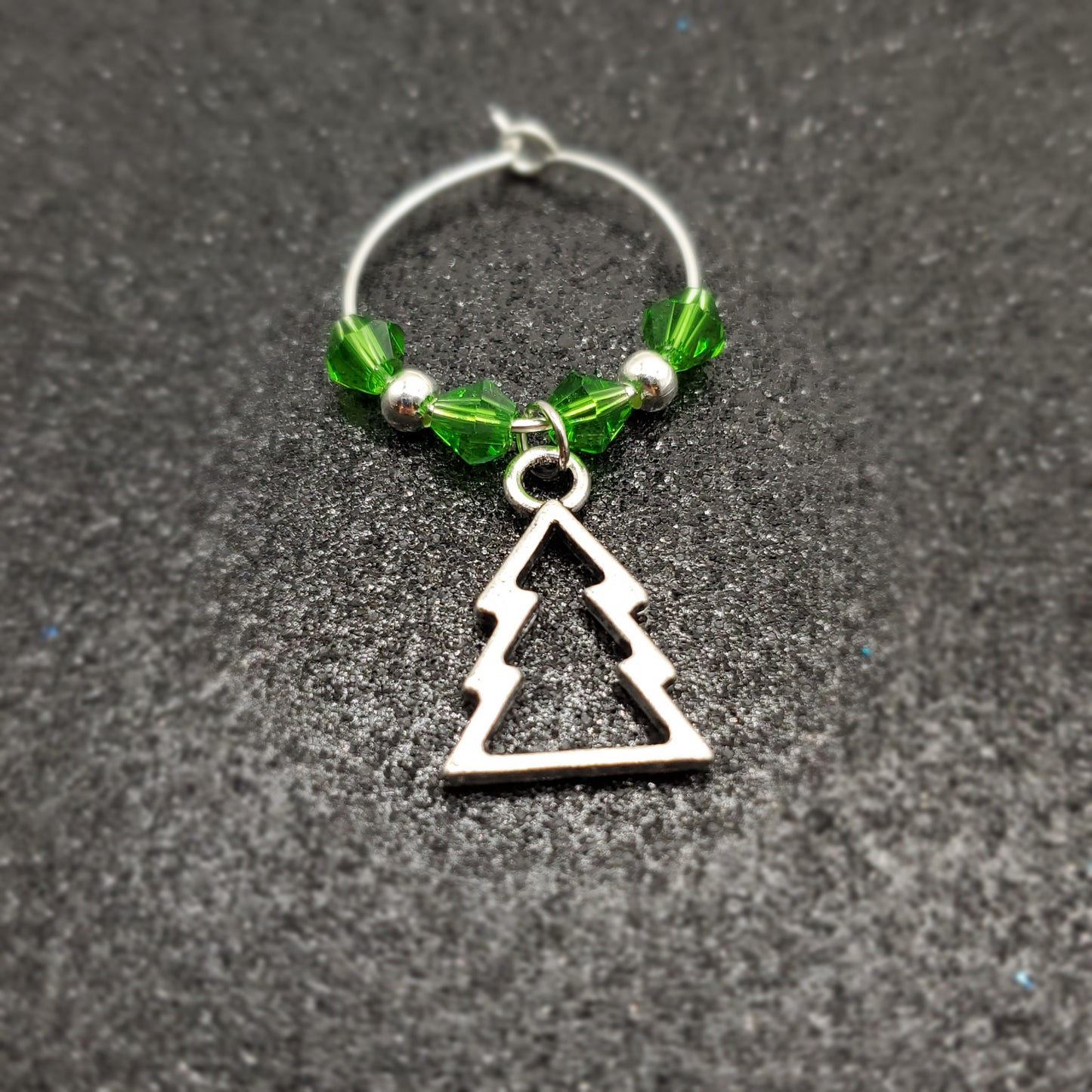 Silver wine glass charm with Christmas tree and green beads on dark surface